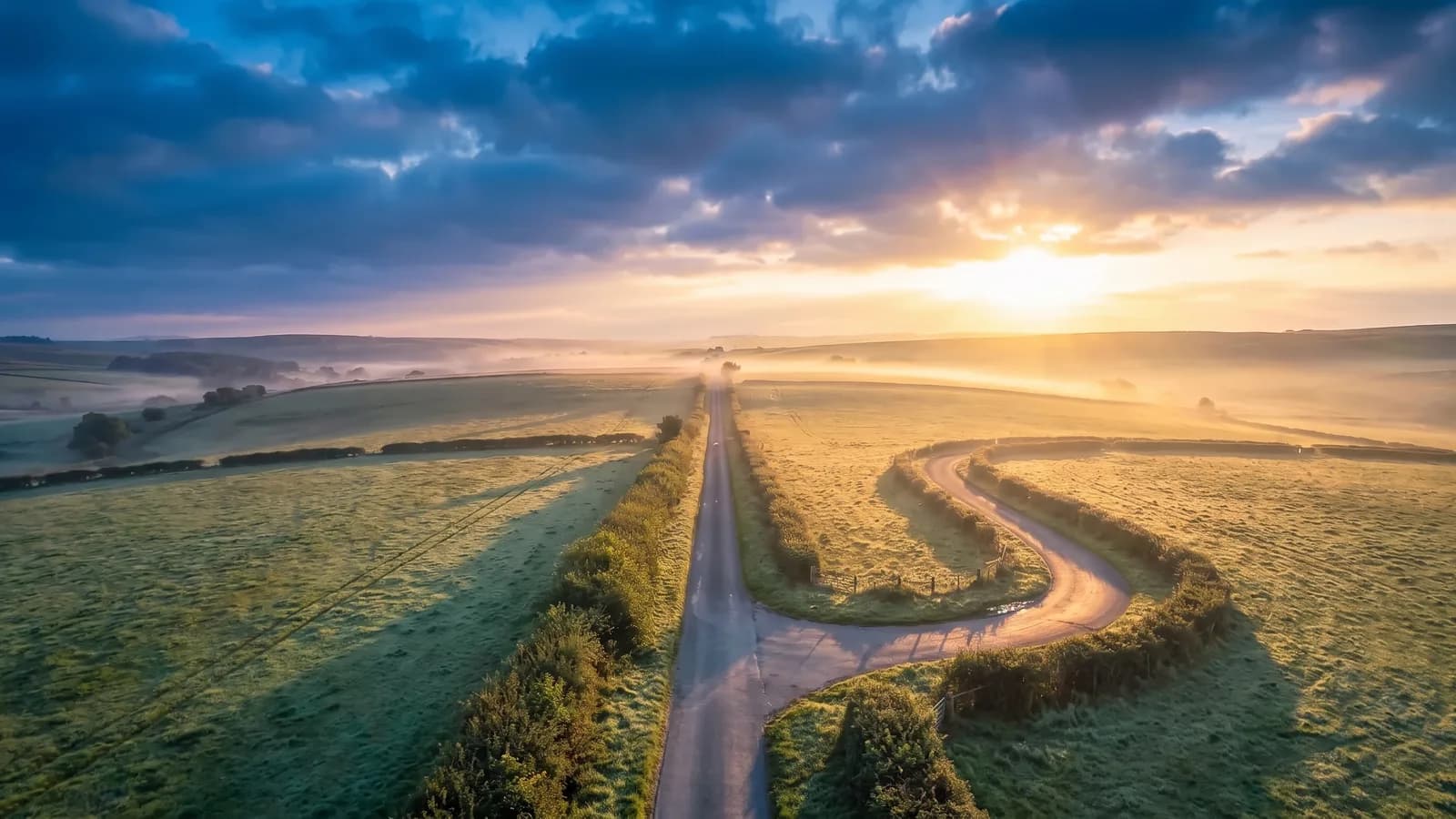 A straight country lane running toward a misty sunrise, with a second lane curving back on itself in a wide U through the dewy fields