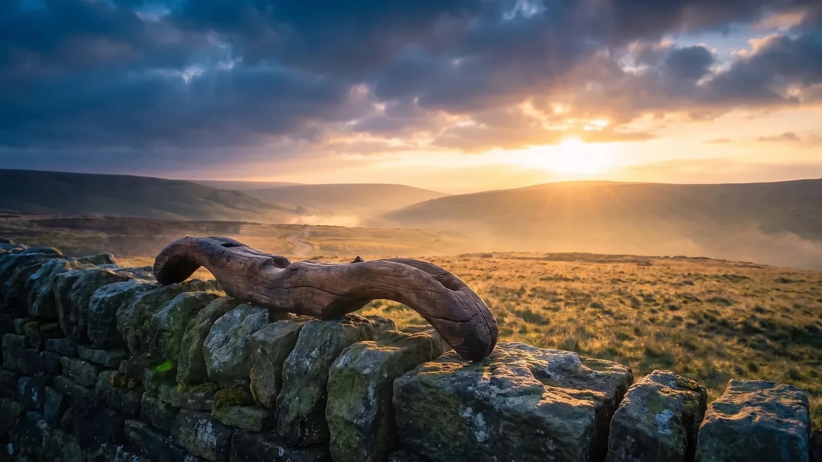 A worn, hand-carved wooden yoke resting on a mossy dry-stone wall, with open moorland and a misty valley beyond under the rising sun